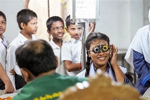 A schoolgirl smiling through a trial frame at a Netram eye camp, her classmates behind her